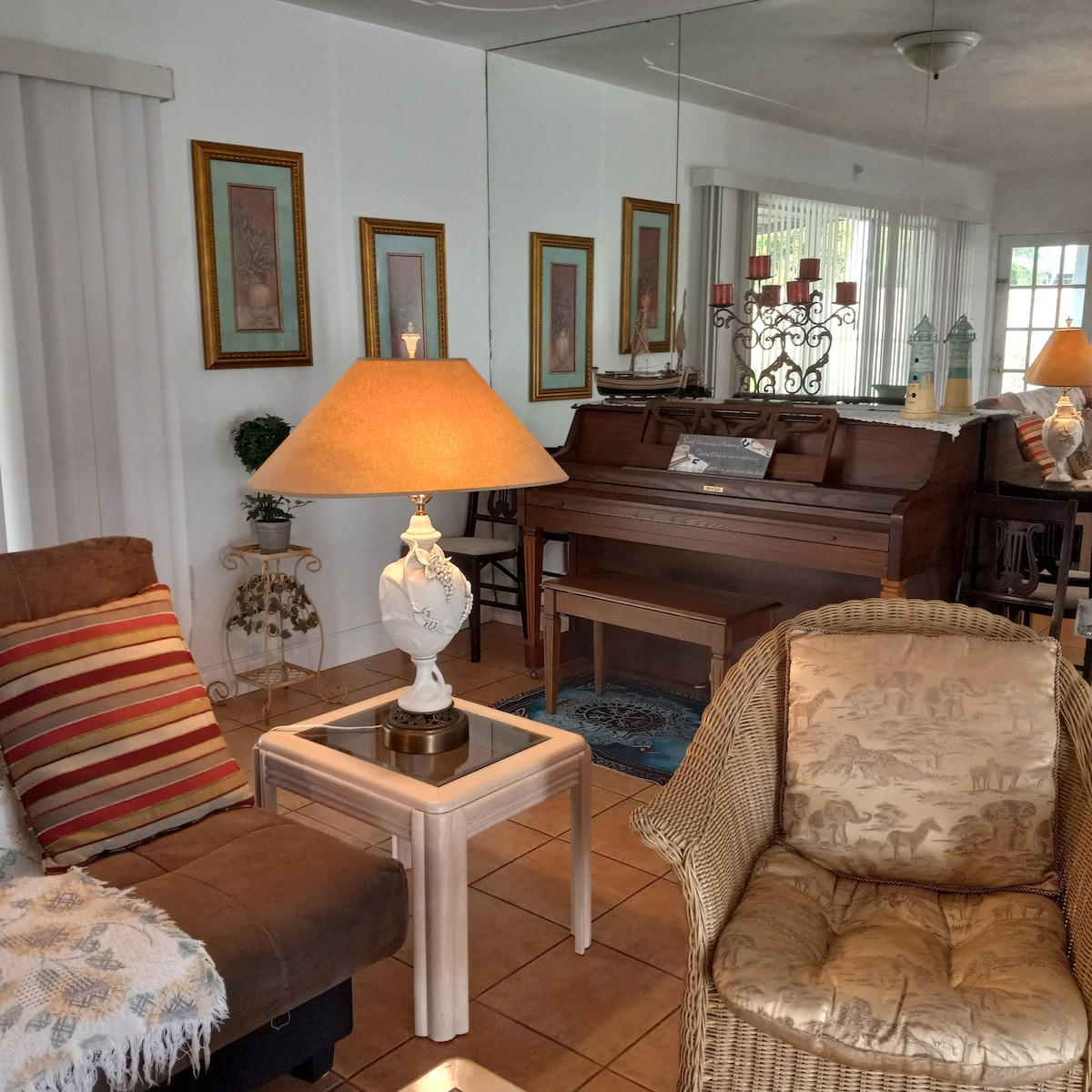 A cozy living area is visible, featuring a mix of comfortable seating including a striped armchair and a wicker chair. A small lamp provides warm light on a side table. Reflections in the mirrors enhance the spaciousness, while a piano is seen in the background.