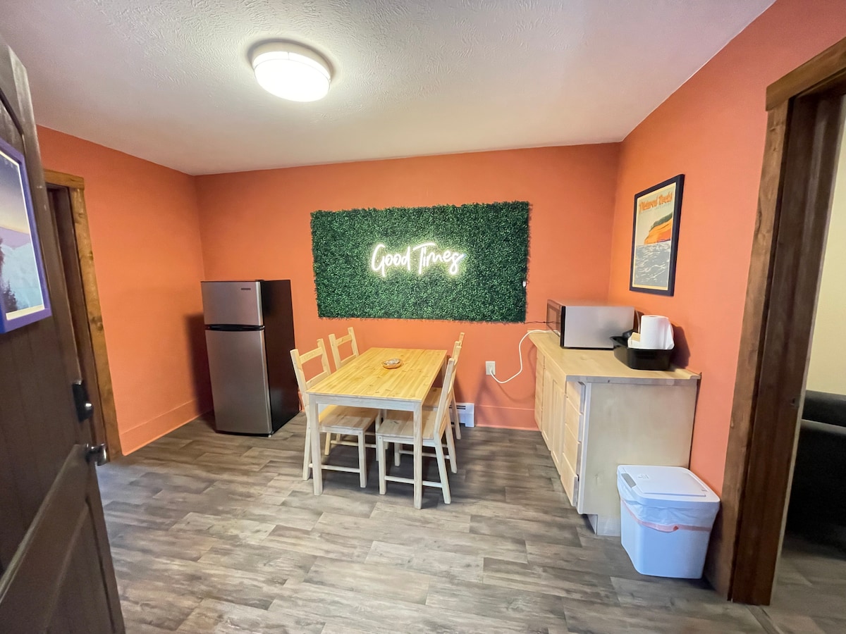 A dining area is highlighted by an orange wall and features a wooden table surrounded by four chairs. A small refrigerator, microwave, and a beige cabinet are positioned against the wall. A green accent wall with the text 'Good Times' adds a cheerful touch to the space.