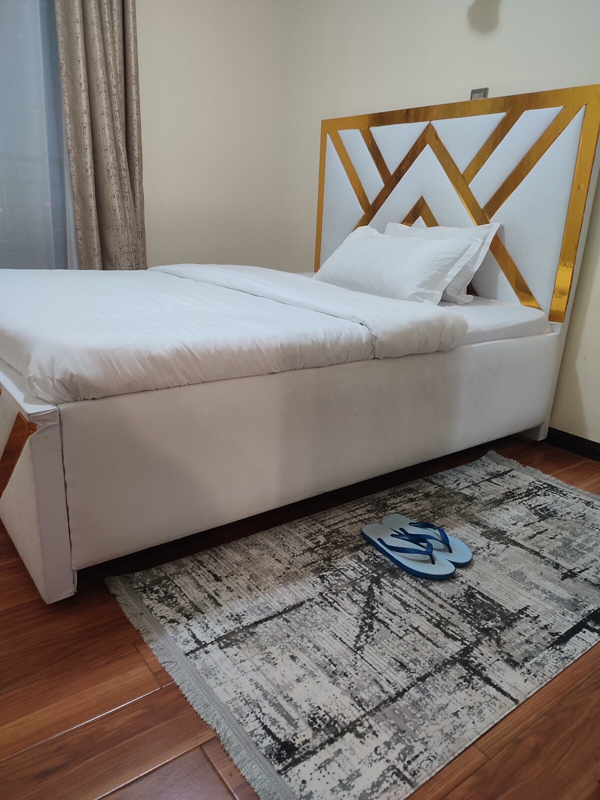 A neatly made bed with crisp, white linens is positioned against a stylish headboard featuring geometric patterns. A soft rug is placed underfoot, displaying a subtle design. A pair of blue slippers lies casually on the rug, completing the comforting atmosphere of the room.