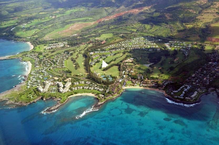 An aerial view displays the lush, green landscape of Kapalua, featuring the Bay Golf Course and nearby beaches. The coastline is marked by vibrant blue waters contrasting with the greenery, while a mix of residential areas and golf facilities can be seen in the distance.