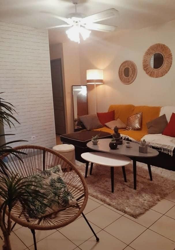 A welcoming living area features a comfortable couch adorned with bright cushions. A coffee table sits at the center, surrounded by two small tables. A decorative plant is positioned in a chair, while soft light casts a warm glow throughout the space.