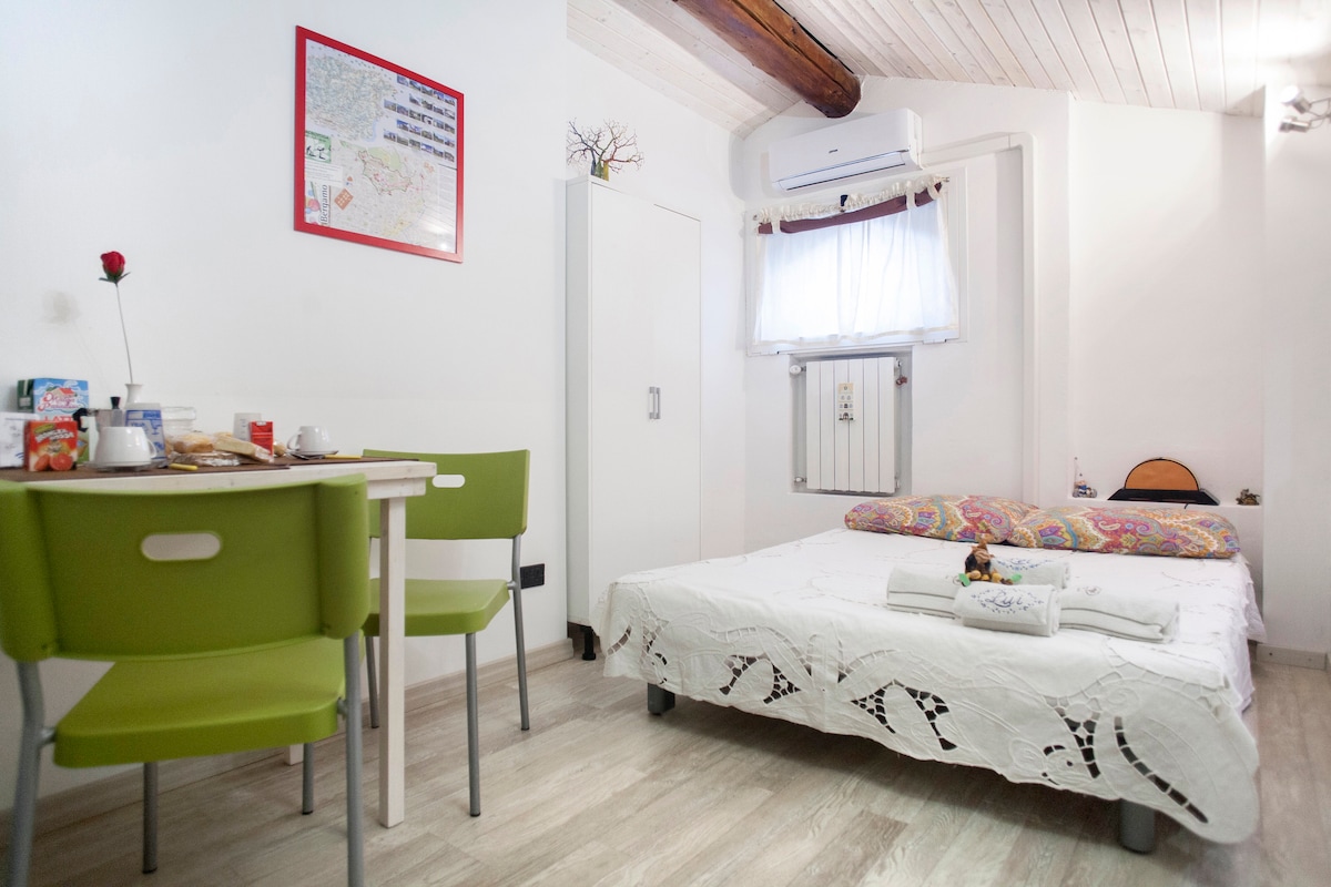 A well-lit room features a spacious bed with a white quilt and colorful pillows. A small dining table with two green chairs is positioned nearby. An air conditioning unit and a white wardrobe are visible, complemented by natural light streaming in through a window with sheer curtains.
