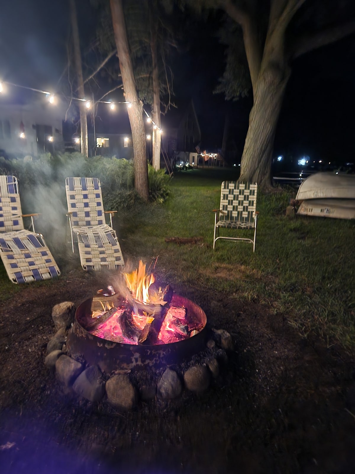An outdoor fire pit is surrounded by stones and features a vibrant fire. Two folding chairs are positioned around the pit, and string lights are visible in the background, casting a soft illumination over the grass and nearby trees.