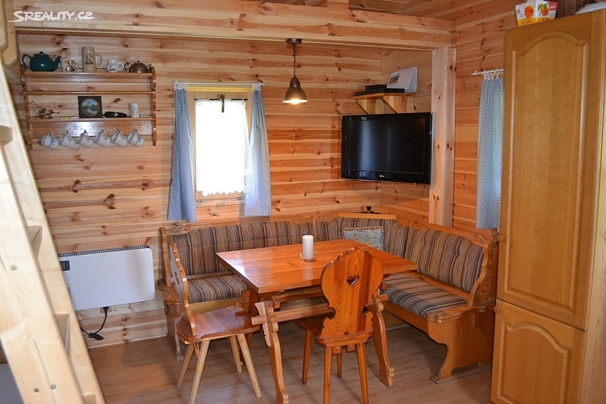 A cozy dining area features a wooden table surrounded by a curved bench and chairs, set against warm wooden walls. A television is mounted above the bench, and soft lighting illuminates the space. Shelves display dishware and decorative items, contributing to a homey feel.