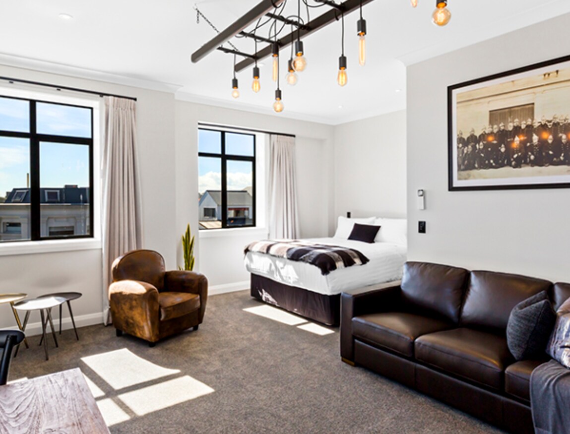 A spacious studio apartment features large windows with city views and natural light. A generous king-size bed is positioned alongside a queen-sized sofa bed. Comfortable seating is provided by an armchair and a leather sofa, while pendant lighting enhances the modern design.