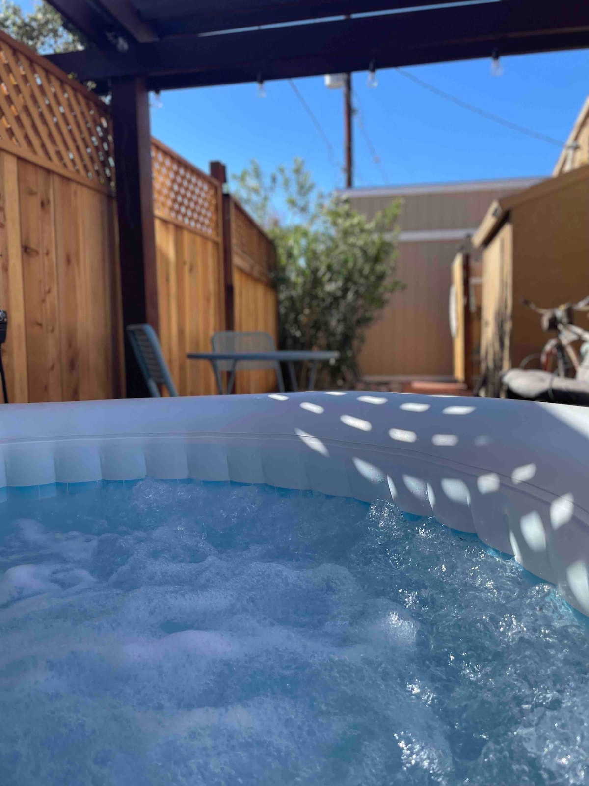 The image captures a close view of a hot tub filled with bubbling water, surrounded by a fenced patio area. A sunny sky is visible above, with shadow patterns cast by a pergola. A small table and chairs can be seen in the background, adding to the outdoor relaxation space.