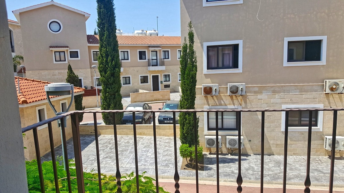 A view from a balcony captures a courtyard area, showcasing stone-paved grounds bordered by greenery. Several air conditioning units are visible on the neighboring buildings, while a parking area contains parked vehicles. Tall cypress trees provide natural accents to the scene.