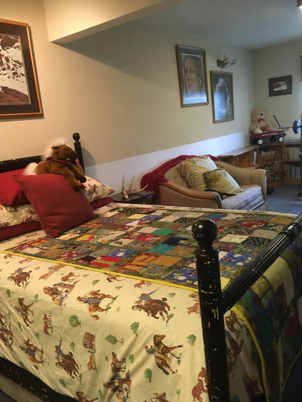 A spacious sleeping area features a bed adorned with a colorful quilt depicting various patterns. Decorative pillows add comfort. Nearby, a seating area is visible with an armchair and additional pillows, while art pieces and wooden accents are displayed on the walls.