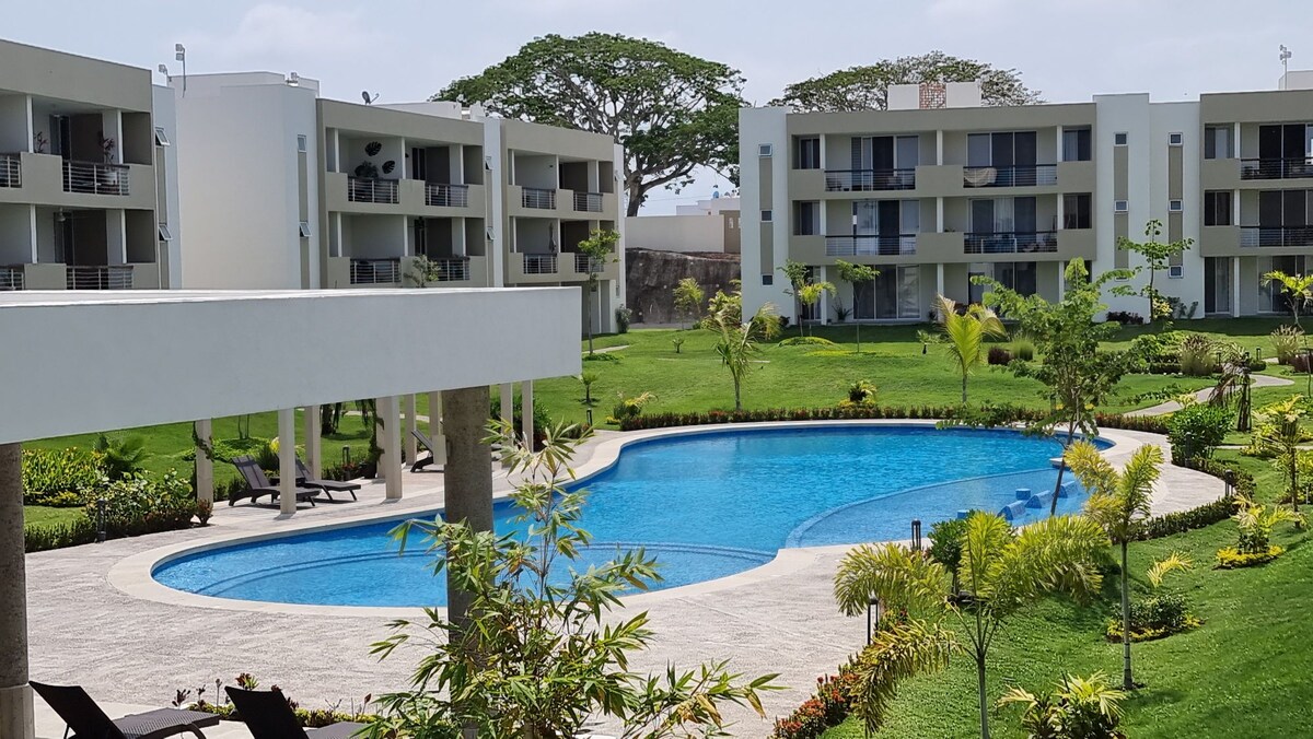 The common area features a swimming pool surrounded by lush green lawns and landscaped gardens. Multiple apartments are visible in the background, offering a view of balconies and outdoor spaces. The calm water reflects the bright sky above, enhancing the serene environment.