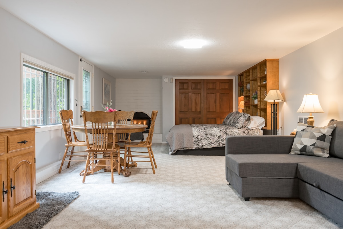 An open living area is highlighted by a cozy sofa with patterned cushions and a spacious bed dressed in soft linens. A wooden dining table with chairs is set near a window, enhancing the light, airy feel of the space. An inviting side cabinet adds functionality.