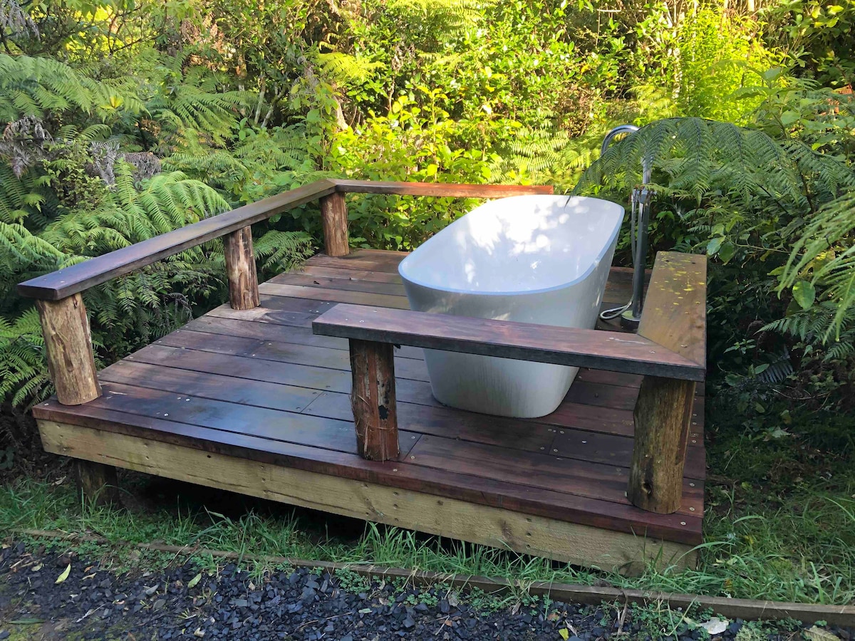 A sleek outdoor bath is positioned on a wooden deck, surrounded by lush ferns and native shrubs. Natural wood accents frame the bath, creating a serene space for relaxation amidst the greenery.