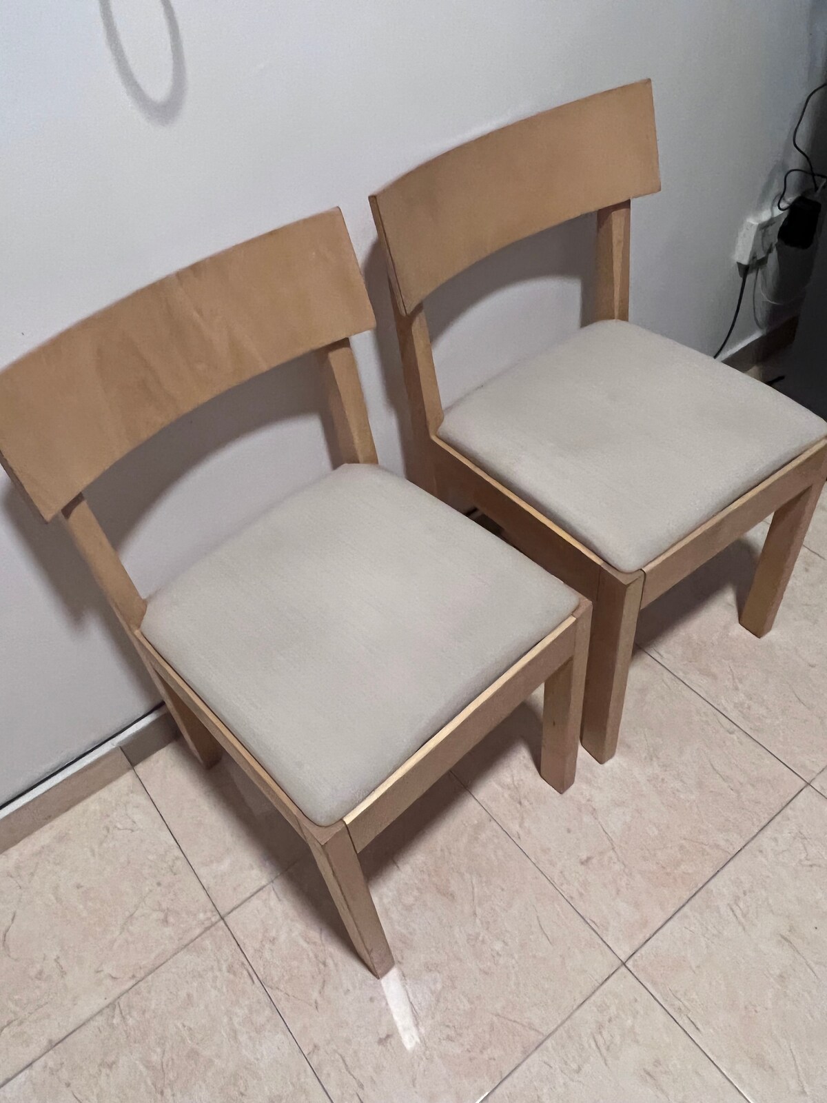 Two wooden chairs are positioned side by side against a light-colored wall. Each chair features a simple design with a flat backrest and beige upholstered seats. The tiled floor reflects subtle light, enhancing the room's calm ambiance.