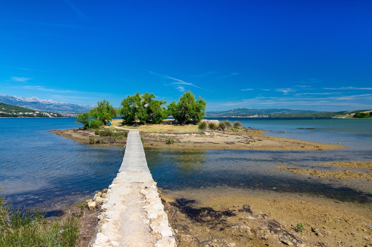 An expansive view of a tranquil bay is captured, featuring a narrow, stone pathway leading to a small island surrounded by calm waters. Lush greenery is present on the island, while mountains are faintly visible in the background under a clear blue sky.