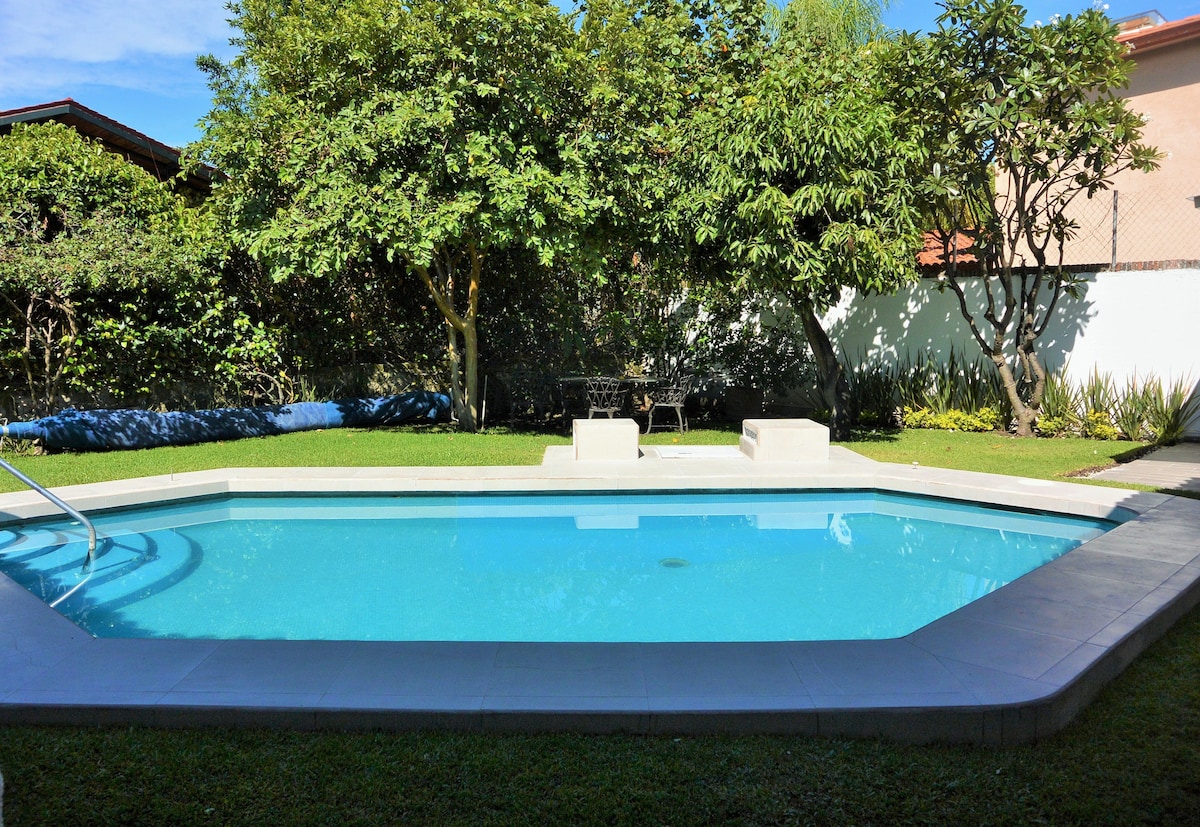 A private pool is surrounded by lush greenery and fruit trees, creating a peaceful outdoor space. Two lounge chairs are positioned at the pool's edge, and the clear water reflects bright daylight, inviting relaxation and enjoyment in a tranquil setting.