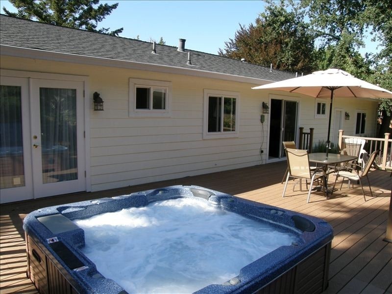 A spacious wooden deck features a hot tub surrounded by bubbles. Comfortable outdoor seating for four is provided under a large umbrella, while a sliding glass door leads into the house. The exterior of the home showcases a simple white façade with multiple windows.