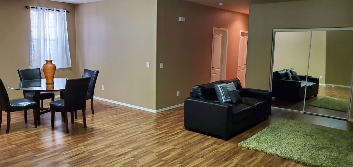 A spacious living area is shown, featuring a dark sofa with a decorative pillow and a green area rug. A dining table with four chairs is set, complemented by a tall vase. Mirrored doors add depth to the room, enhancing the overall open feel.