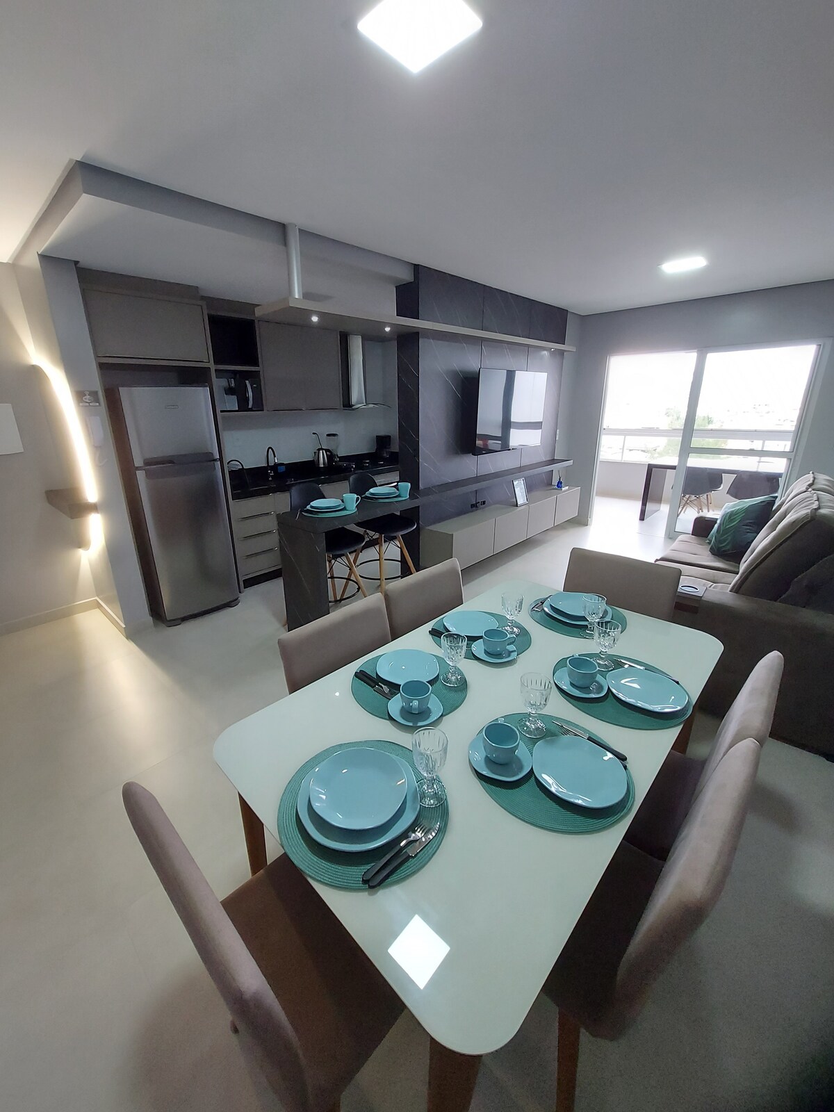 An open kitchen and living area are visible, featuring a modern dining table set for six. Turquoise dishware and glassware are neatly arranged. The kitchen includes sleek cabinetry and appliances. A comfortable seating area with soft furnishings can be seen in the background, with natural light coming through the window.