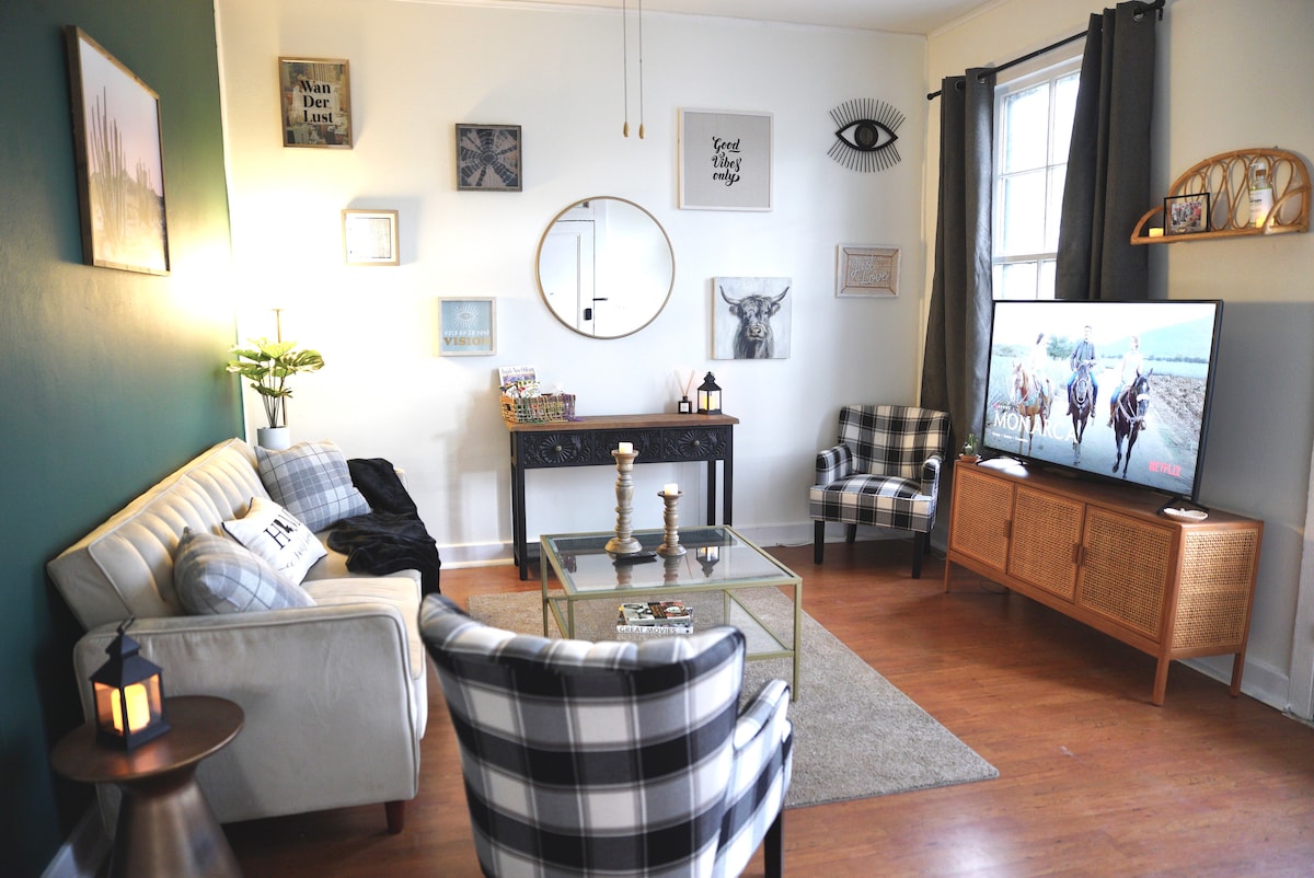 A comfortable living area is presented with a mix of modern and rustic decor. A large television occupies one corner, while a glass coffee table is centered on a plush area rug. A combination of seating includes a neutral-toned couch and plaid chairs beside a decorative console.