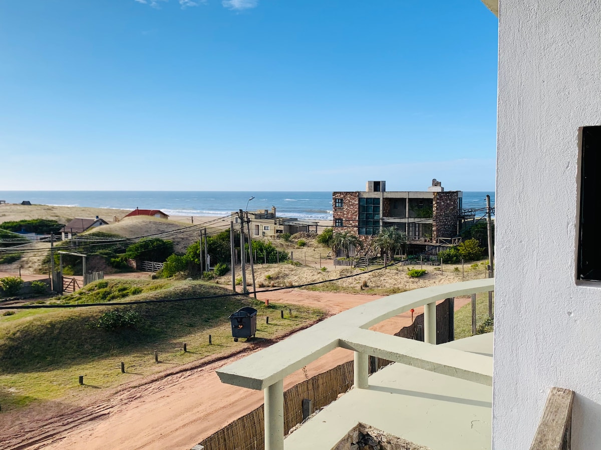 A view from a balcony overlooks a sandy expanse leading to the beach. The ocean glistens under clear blue skies, with a gentle slope of greenery in the foreground and nearby buildings adding to the coastal scenery.