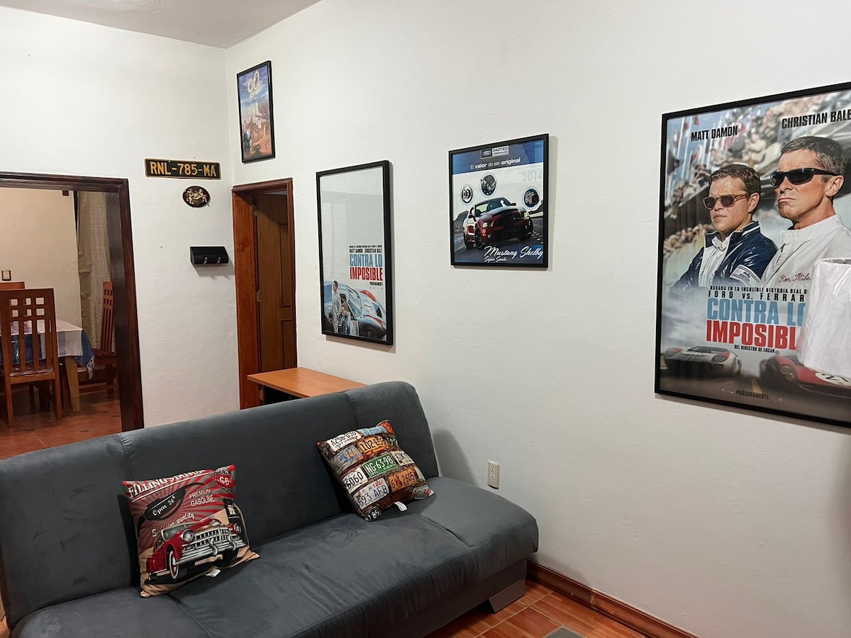 A living area is displayed, featuring a gray sofa adorned with colorful cushions. Two framed posters and a mirror are mounted on the light-colored walls. A wooden dining table and chairs are visible in the background, contributing to the room's functionality.