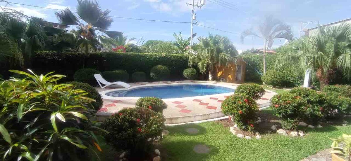 A serene outdoor space features a private swimming pool surrounded by well-manicured gardens and tropical foliage. Lounge chairs are positioned beside the pool, while decorative stone pathways meander through the lush greenery.