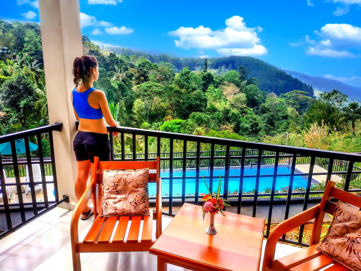 A private balcony is depicted with a wooden table and two chairs. A flower vase rests on the table. The view showcases lush greenery and rolling hills, while a swimming pool is visible in the distance under a clear blue sky.