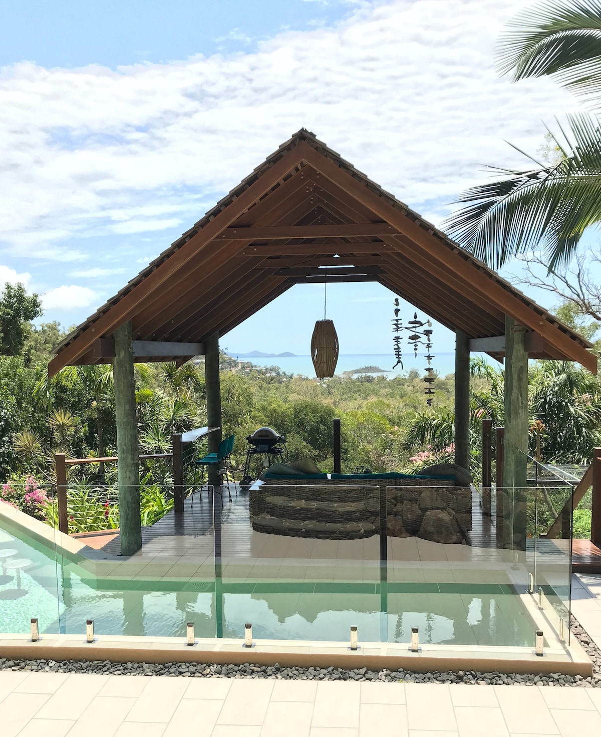 A spacious outdoor pool hut is framed by tropical plants, featuring a stone seating area and wooden beams overhead. The area provides views of the surrounding landscape, where palm trees and green hills contrast with the blue sky. Soft lighting is positioned along the pool's edge.