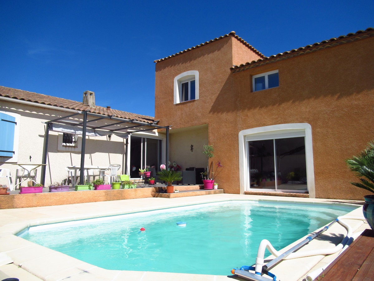 The exterior of the property is highlighted by a private pool surrounded by colorful flowerpots. A spacious terrace is visible with seating arrangements and a covered area adjacent to the house. Sunlight reflects off the pool water under a clear blue sky.