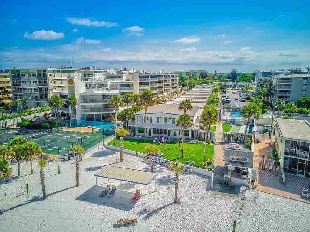An aerial view captures a serene beachside community, featuring palm trees and sandy areas leading to the beach. Nearby, tennis courts and landscaped spaces are visible, along with multiple buildings designed for guest accommodations, highlighting access to outdoor relaxation areas.