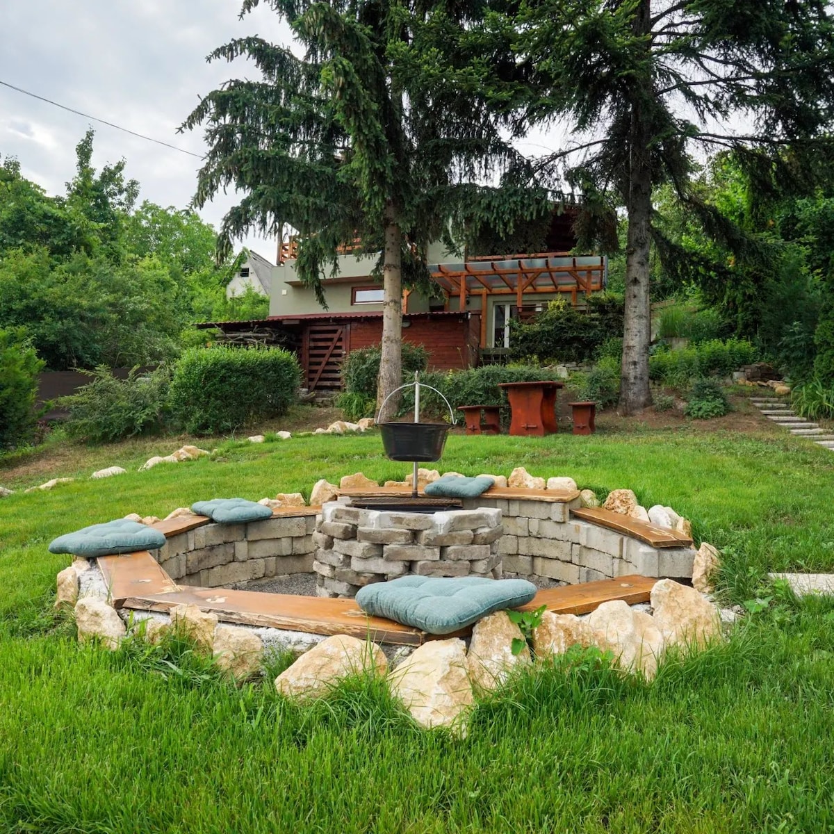 A fire pit area is surrounded by stone and wooden seating, inviting relaxation. Cushions are placed for comfort. Lush green grass and a variety of trees frame the scene, with a house nestled in the background, enhancing the serene outdoor environment.