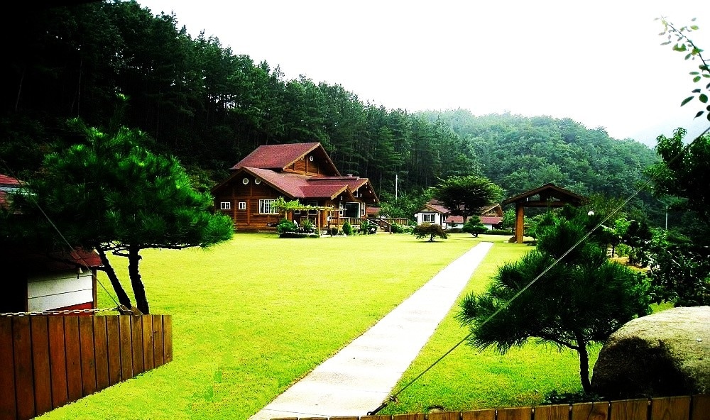 A spacious lawn is surrounded by trees and leads to a two-story log house, characterized by a red roof. A traditional pavilion is visible, adding to the serene landscape. The area is well-maintained, evoking a sense of privacy and tranquility.
