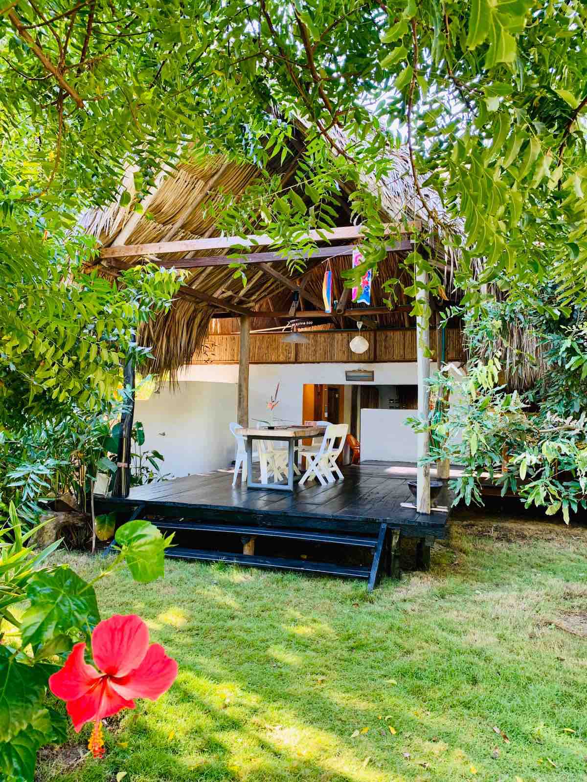 A tropical cabin is nestled among lush greenery, featuring a thatched roof and a wooden porch. A dining table with white chairs is seen on the patio, surrounded by vibrant plants and colorful flowers, creating a serene and natural atmosphere.