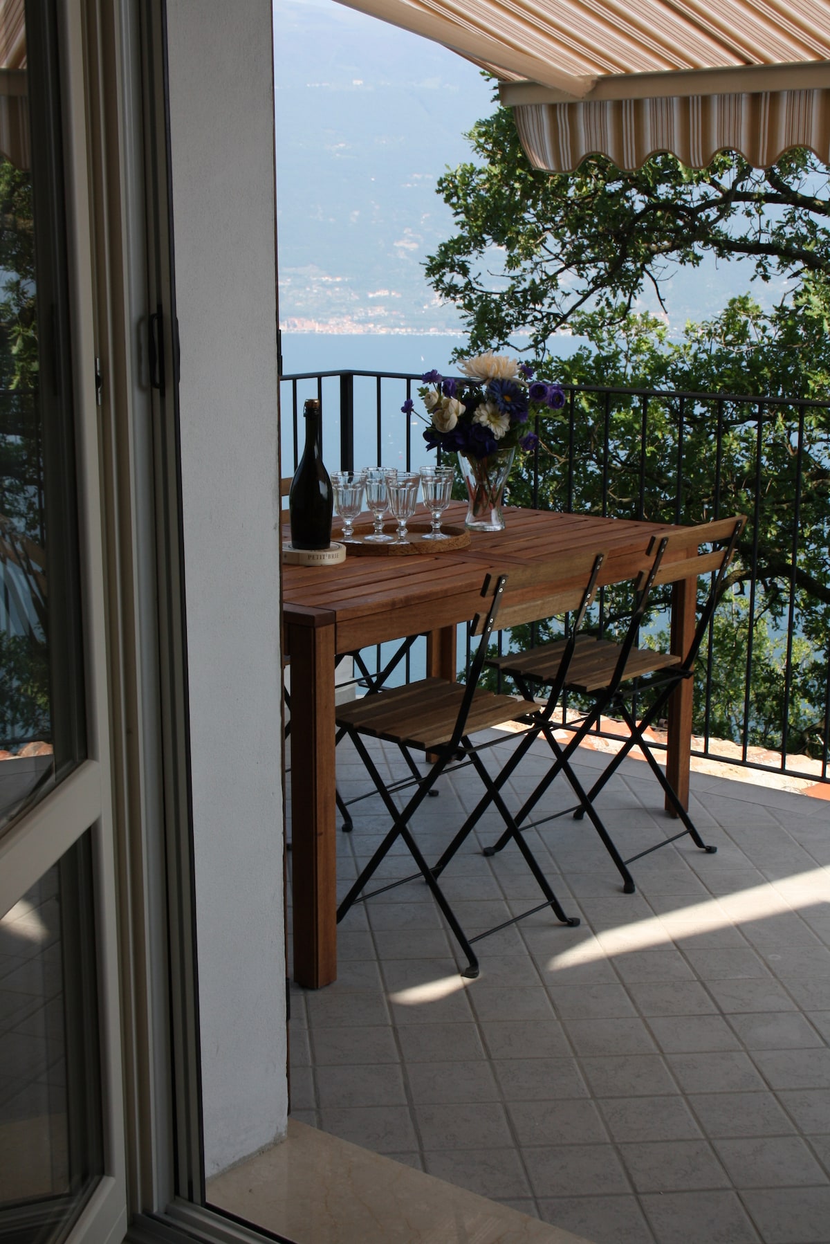 An open balcony features a wooden dining table set for four, accompanied by two folding chairs. A vase of vibrant flowers sits at the center. The balcony is shaded by a striped awning, with views of the surrounding greenery and distant lake visible beyond the railing.