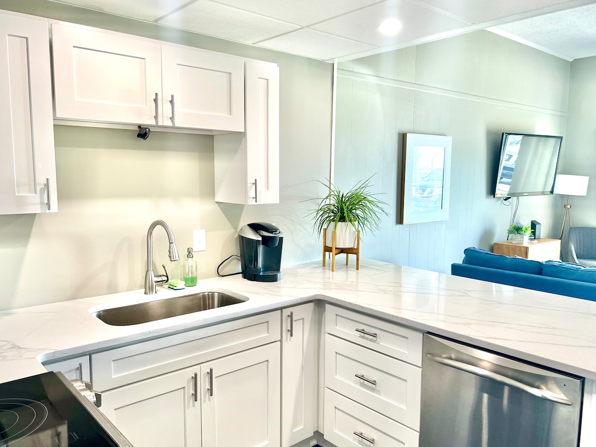 The newly renovated kitchen features sleek, white cabinetry and modern appliances. A stainless steel dishwasher and a stainless steel sink are present, complemented by a marble countertop. A coffee maker and decorative plant enhance the space, with a glimpse of the living area visible in the background.