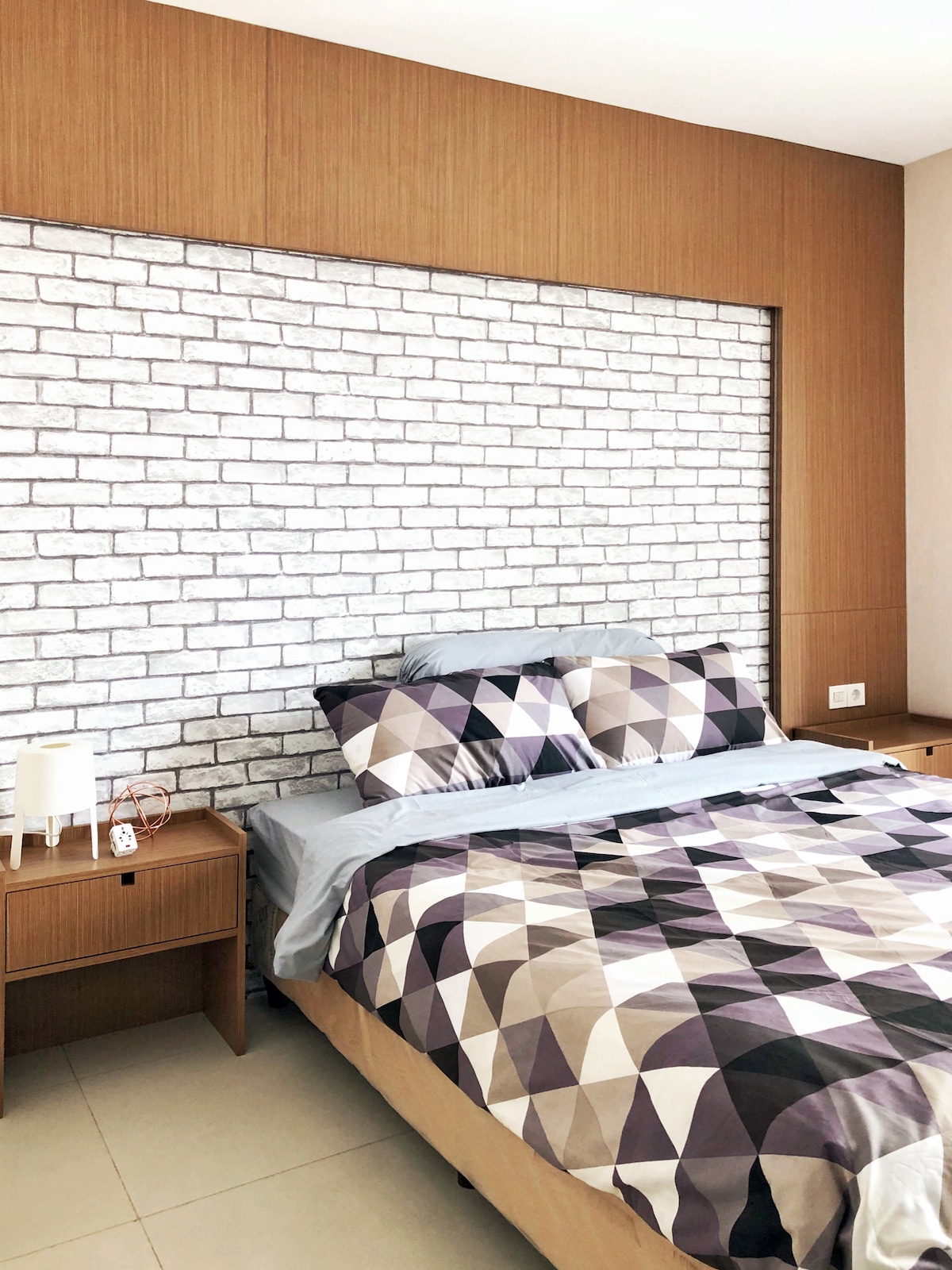 A cozy bedroom is highlighted by a patterned bedspread in shades of gray and purple. A headboard of wooden panels complements a decorative brick wall. Two bedside tables are positioned on either side of the bed, enhancing the functional and clean design.