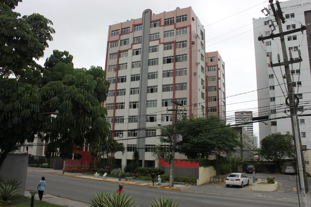 A residential building is prominently displayed with multiple floors and large windows. The exterior features a combination of light and dark hues, along with balconies. Surrounding greenery includes tall trees and shrubs, enhancing the building's urban setting. Street-level views showcase nearby infrastructure.