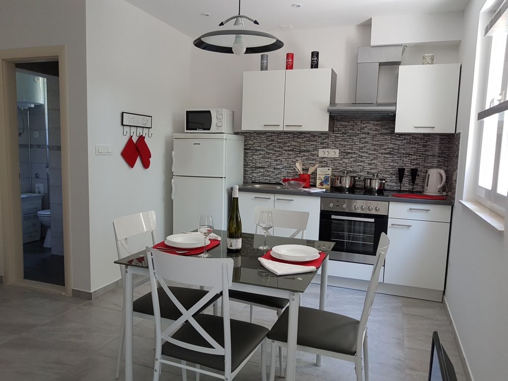 A modern kitchen and dining area feature a glass-top table set for four, with white chairs and red placemats. A full-size fridge and microwave are visible, alongside a stainless-steel oven and stovetop. Light enters from a window, illuminating the neutral-toned cabinetry.