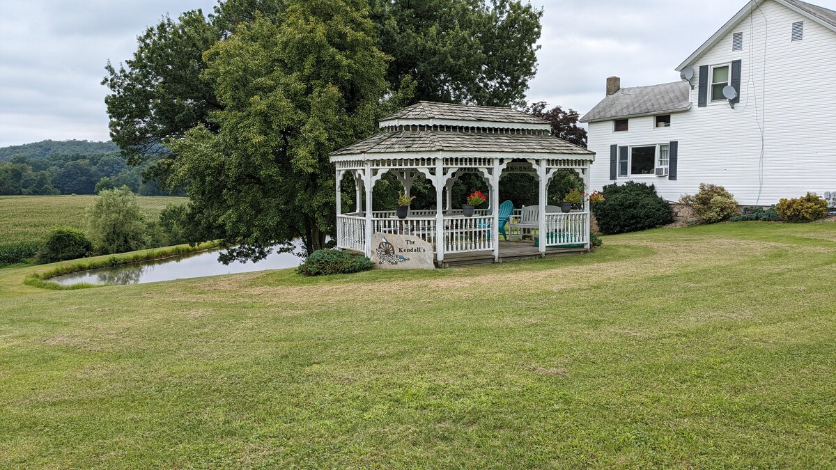 A wooden gazebo with a slatted roof stands in an open grassy area, surrounded by greenery. Colorful potted plants are positioned at the entrance, and a calm pond can be seen in the background, framed by gentle hills and trees.