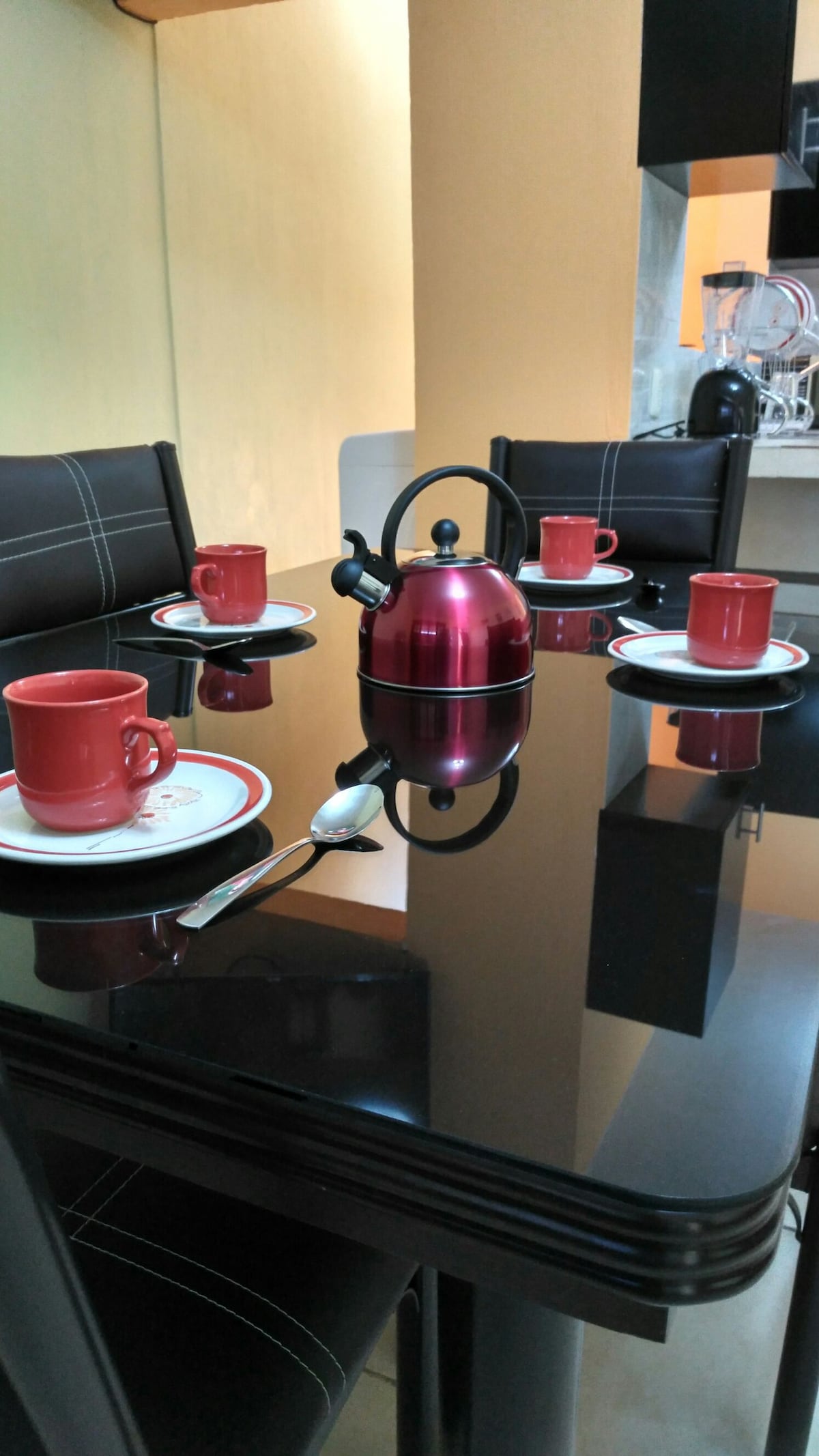 A modern dining table is set with four red cups and matching plates, arranged around a reflective black tabletop. A kettle in a deep red hue sits at the center, accompanied by a silver spoon, while soft light enhances the warm ambiance of the space.