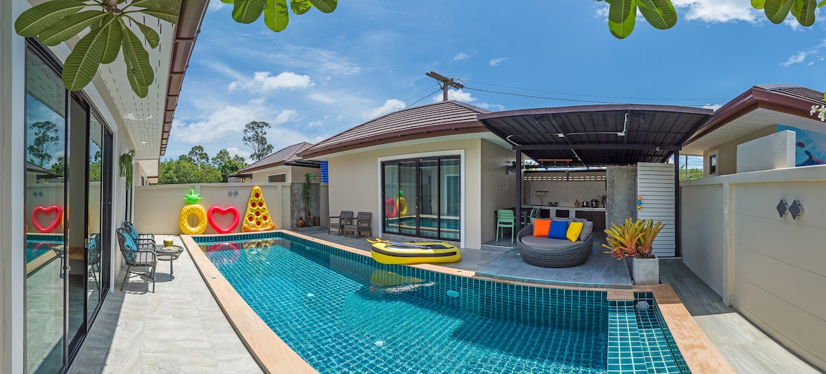 An inviting outdoor space features a vibrant blue swimming pool surrounded by sun loungers and playful floatation devices. Large glass doors provide views into the villa, while tropical plants add a lush touch to the pool area, illuminated by natural sunlight.