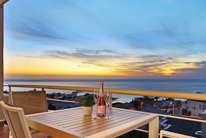 A private balcony features a table set for two, with a bottle of sparkling wine and glasses. The ocean horizon is visible at sunset, showcasing a blend of warm colors in the sky, while a succulent plant adds a touch of greenery.