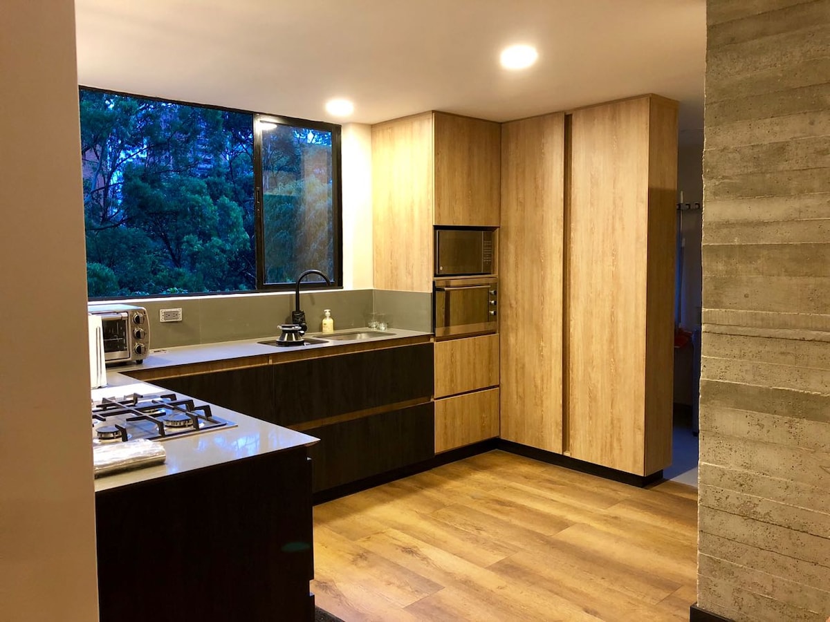 A modern kitchen is featured with wooden cabinetry and sleek stone countertops. A large window offers natural light and views of greenery outside. Appliances such as a stove, microwave, and oven are included. Warm wood flooring complements the contemporary design.