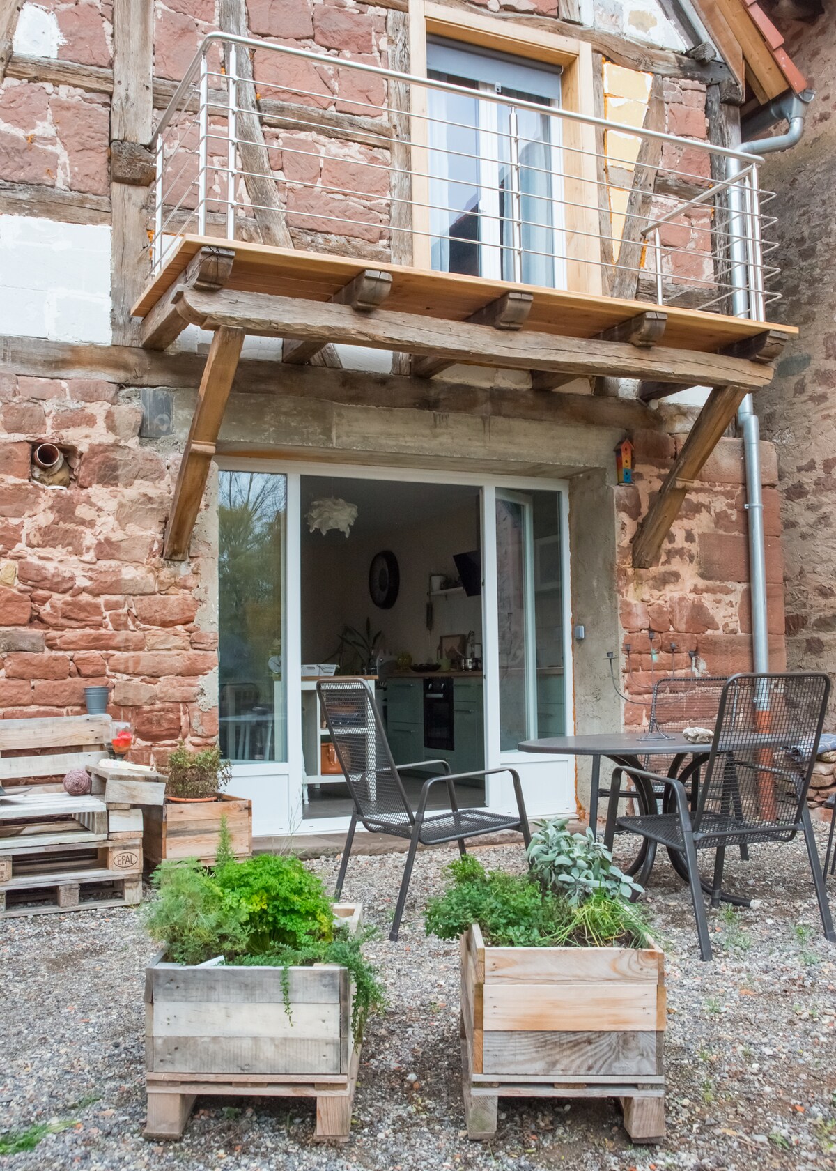 A charming outdoor view features a wooden balcony above glass doors, leading into a welcoming interior. Below, two wooden planter boxes filled with greenery are positioned beside a small seating area with black chairs. The rustic stone walls contribute to the overall inviting ambiance.