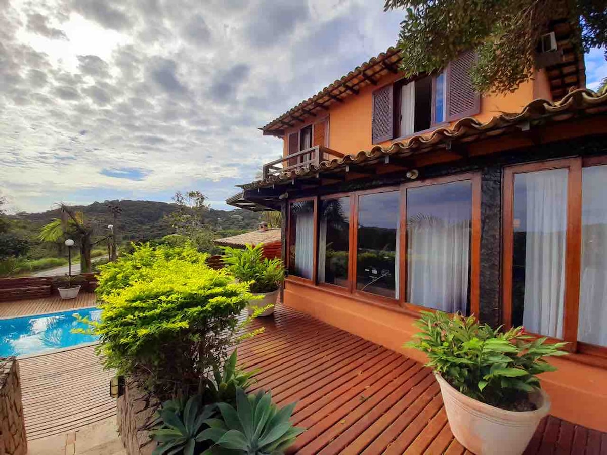 A two-story house is framed by lush greenery and wooden decking. Large windows reflect the natural surroundings, while a covered balcony extends from the upper floor. A pool is visible nearby, inviting relaxation amid the tranquil landscape.