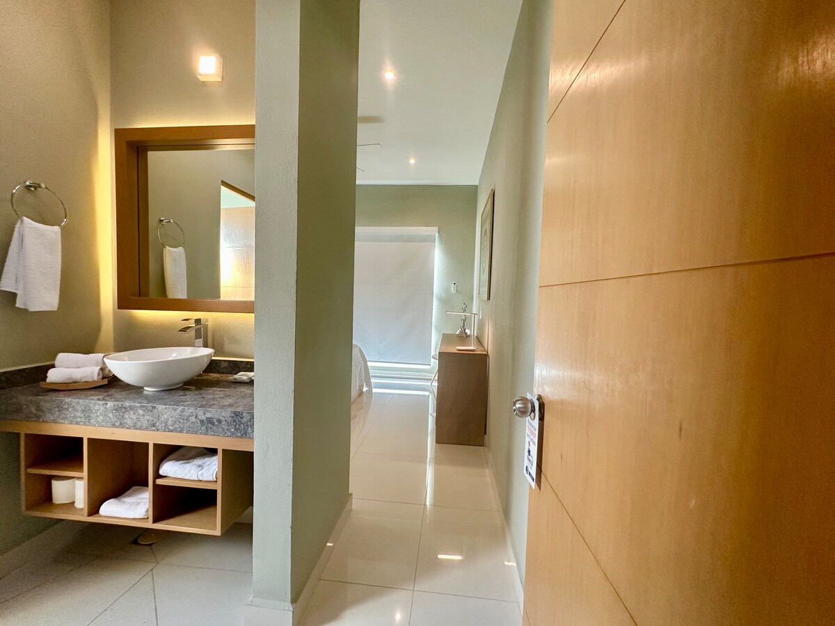 A well-lit bathroom is seen with modern fixtures and a large mirror above the stylish sink. The gray countertop is complemented by wooden cabinetry storing neatly arranged towels. Natural light filters in, enhancing the fresh ambiance of the space.