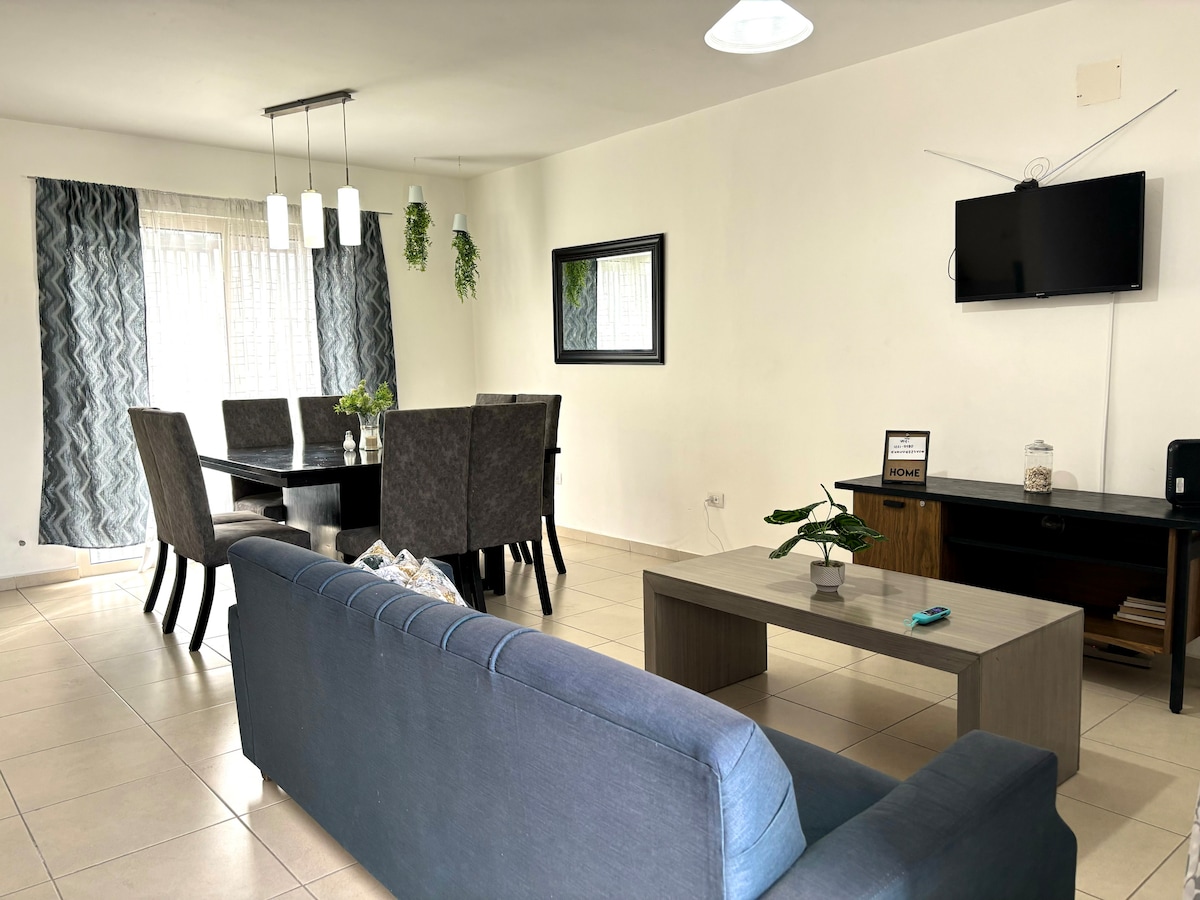An open living and dining area is showcased, featuring a modern sofa in soft blue and a wooden coffee table. A large dining table with six upholstered chairs is positioned under pendant lighting. Natural light filters through sheer curtains, highlighting the spaciousness.