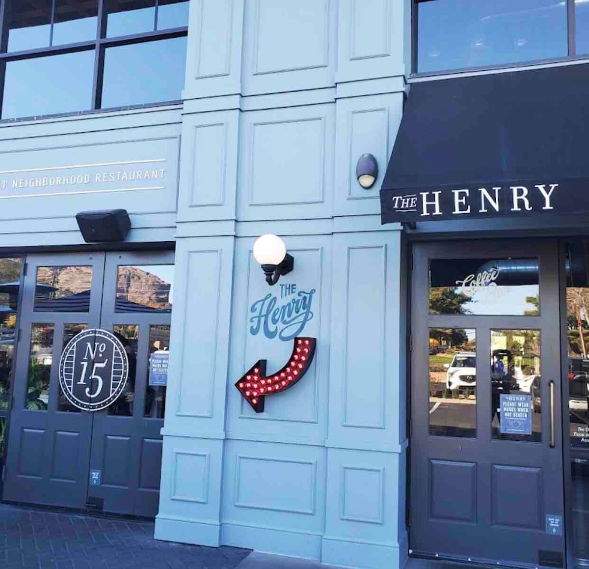 An inviting exterior of a neighborhood restaurant features a stylized sign reading 'The Henry' with red marquee lights. A prominent arrow points towards the entrance, framed by elegant blue paneling. Nearby, decorative circular signage adds to the charming appeal of the facade.