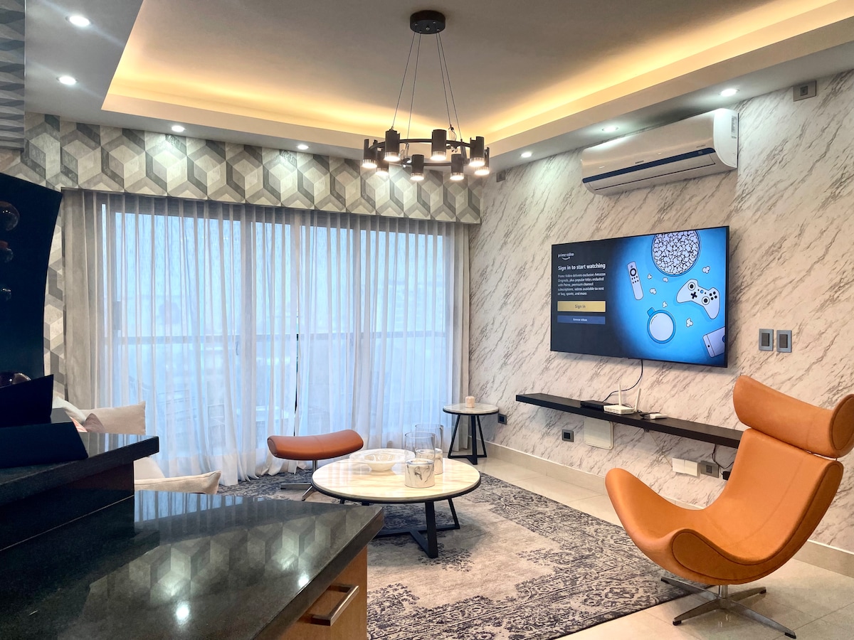 A contemporary living area features a modern seating arrangement with an orange accent chair and two light-colored armchairs around a circular coffee table. A large wall-mounted television is visible, with soft lighting and patterned curtains enhancing the inviting atmosphere.
