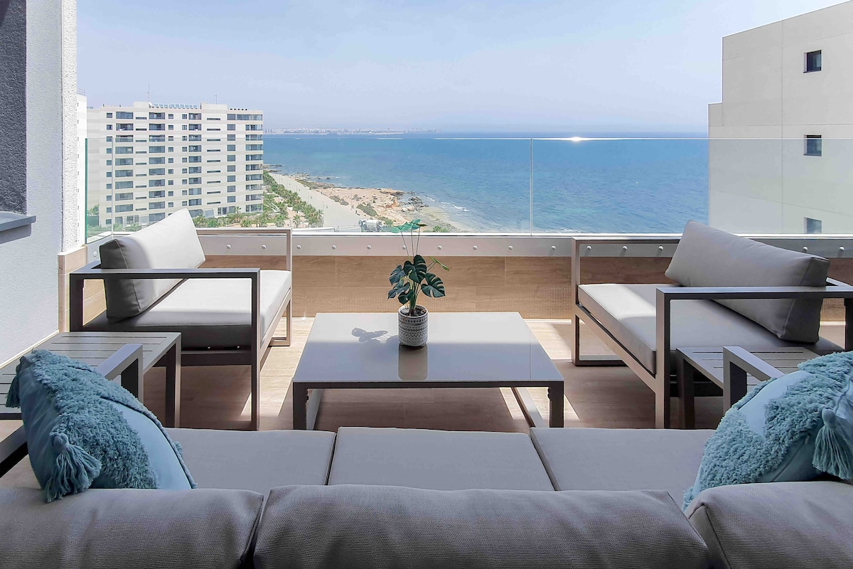 A spacious outdoor seating area is presented, featuring a light-colored sofa and armchairs arranged around a modern coffee table. A potted plant adds a touch of greenery. Behind, expansive glass panels offer a clear view of the beach and ocean.