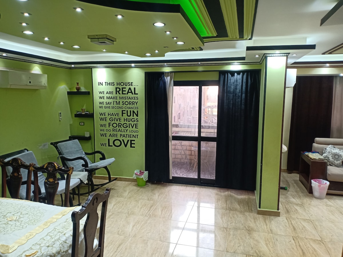 A living area features green-accented walls and a textured ceiling with lighting fixtures. A dining table with chairs is positioned beside a wall decorated with positive affirmations. Large windows with black curtains allow natural light, while a comfortable seating arrangement is visible in the corner.